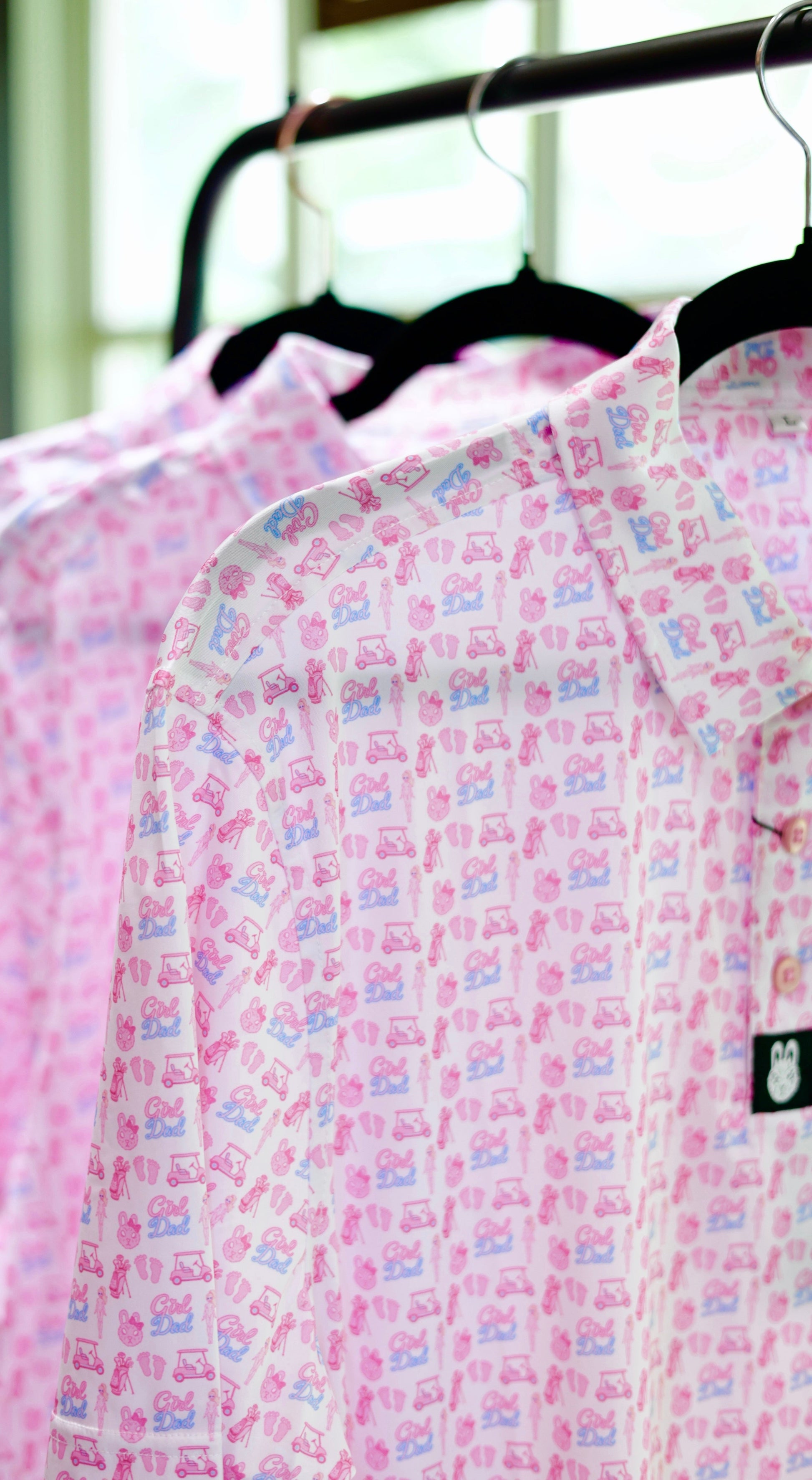 The Girl Dad Polo hanging on a rack featuring a playful pink and blue all-over print on a lightweight performance golf polo.