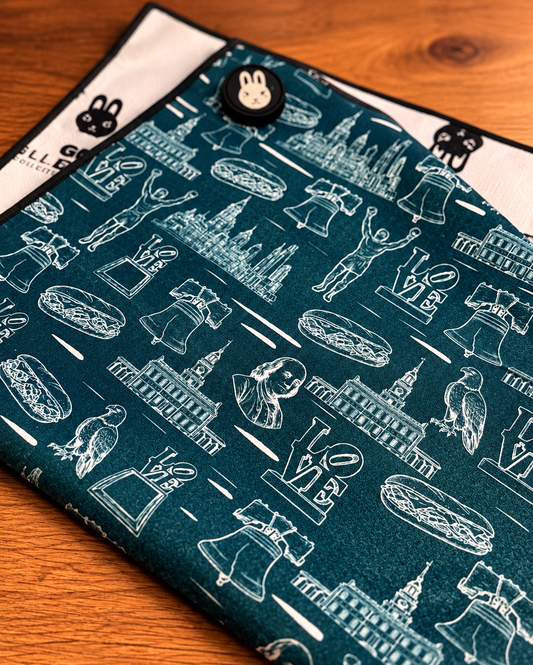 Golf towel The Philly Heritage Towel in dark teal fabric, covered with white line-art Philly landmarks, cheesesteaks, and LOVE symbols, with a black trim and clip.